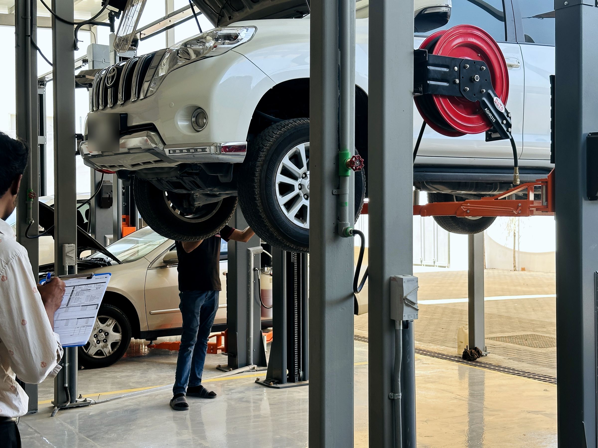 Staff member filling in a vehicle inspection report while a Toyota Prado is checked on a two-post lift